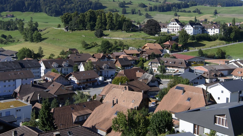 Vogelansicht der Gemeinde Riggisberg. Eine Ansammlung von Häusern mit roten Dächern, eingebettet in grüne Hügel.