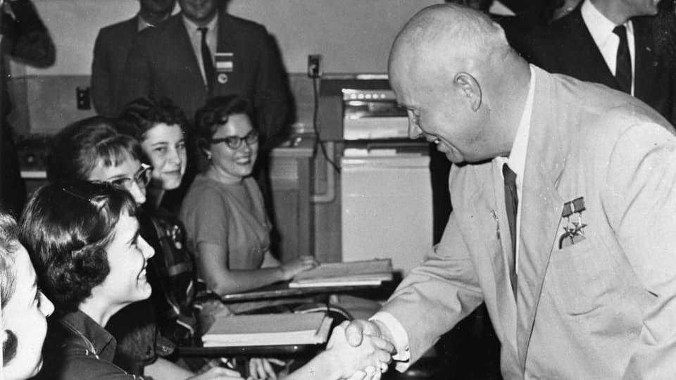 Nikita Chruschtschow schüttelt die Hand einer Frau, die sitzt. Beide grinsen.   