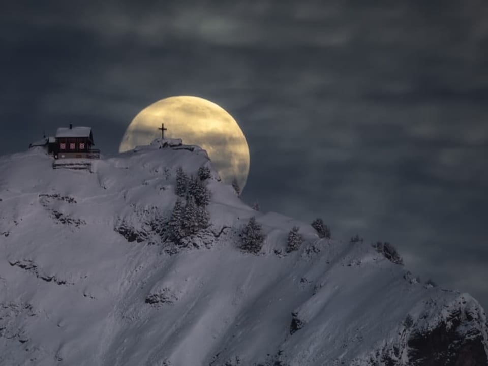 A mountain with a hut and snow behind the full moon glows in the dark.  Clouds high in the sky. 