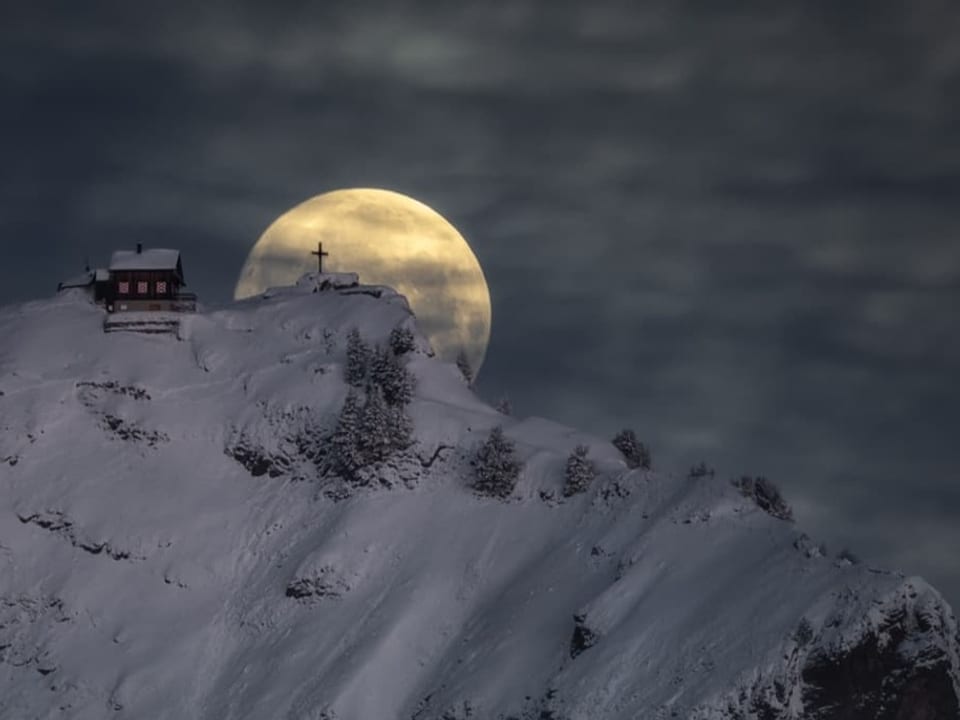 Berg mit Hütte und Schnee, dahinter Vollmond, der in der Dunkelheit scheint. Am Himmel hohe Wolken. 