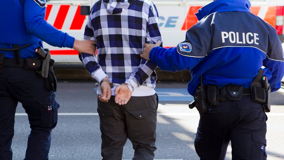 Zwei Polizisten führen einen Mann ab. Seine Hände sind auf dem Rücken zusammengebunden.