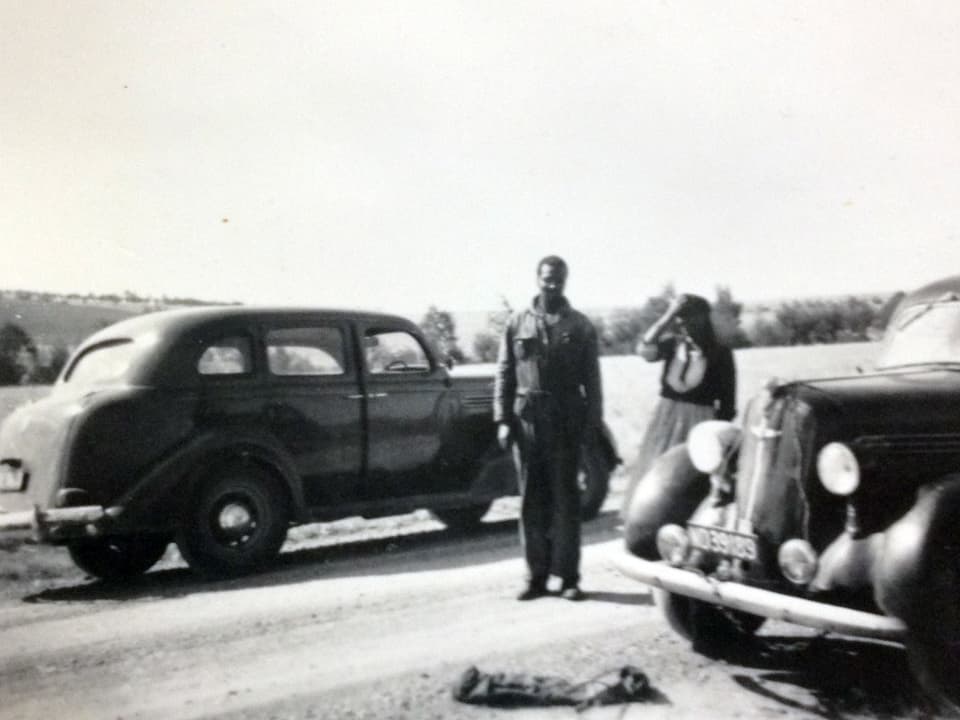 Ein Mann und eine Frau stehen zwischen zwei Fahrzeugen, aus heutiger Sicht Oldtimer.