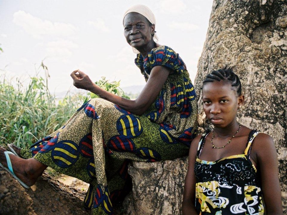 Eine ältere Afrikanerin sitzt an einem Baum, neben ihr steht ein junges Mädchen, das nüchtern in die Kamera blickt.