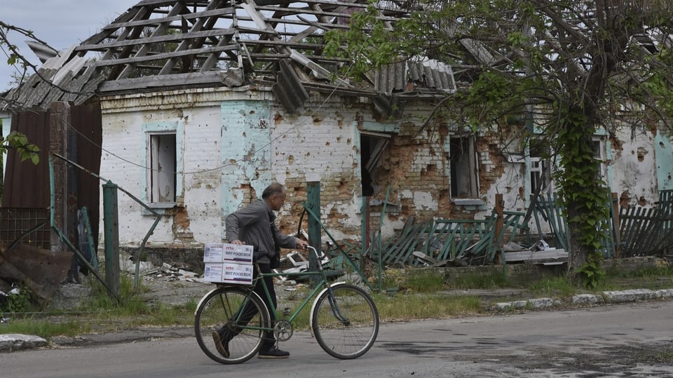 Eine Person mit einem Fahrrad, dahinter ein zerstörtes Haus. 