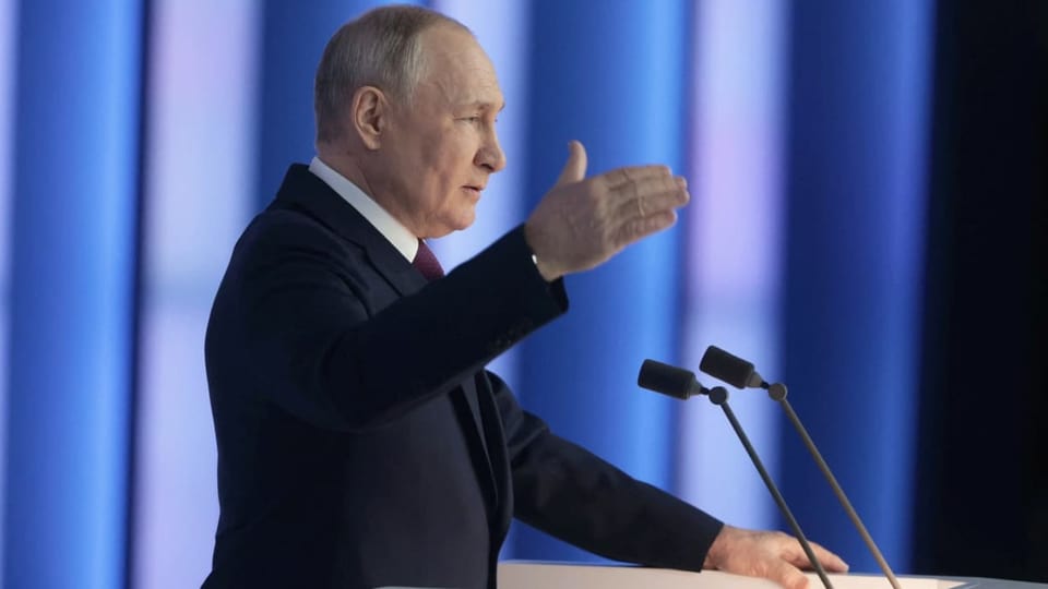 Putin an einem Rednerpult die rechte Hand hebend.
