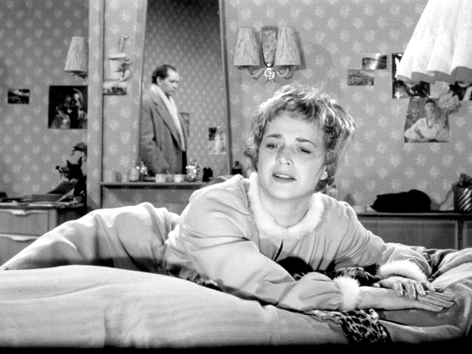 Eine blonde Frau mit kurzen Haaren und einem hellen Mantelkleid mit Pelzkragen liegt auf dem Bett und weint. Der Spiegel im Bildhintergrund verrät die Anwesenheit eines Mannes.