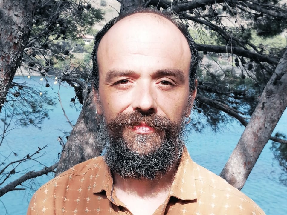 Porträt-Aufnahme eines Mannes mit Bart, in beiden Ohren ein silberner Ring, im Hintergrund Bäume und ein Gewässer.