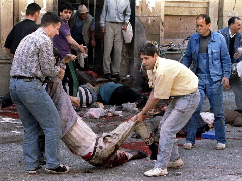 Zwei Männer schleppen eine angeschossene, blutende Person in Sarajevo über den Boden. 