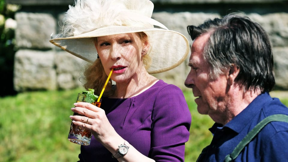 Szene draussen: Eine Frau mit grossem Hut trinkt einen Drink, ein Mann steht daneben.