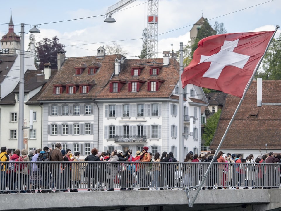 Blick auf eine Brücke in Luzern. Viele Menschen gehen darauf. Im Vordergrund ist eine Schweizer Flagge zu sehen.