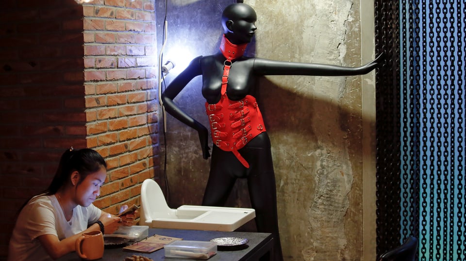 Eine Frau sitzt an einem Café-Tisch, im Hintergrund ein Mannequin mit SM-Outfit.