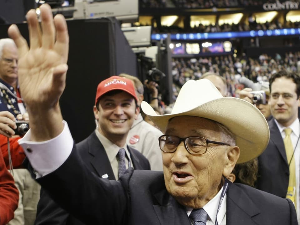 Henry Kissinger trägt eine Brille und einen Hut. Er winkt und lacht.