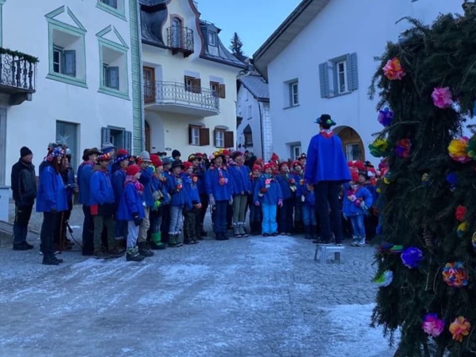 Eine Schulklasse steht singend vor traditionellen engadinger Häusern. Rechts sieht man den Blumenwagen.