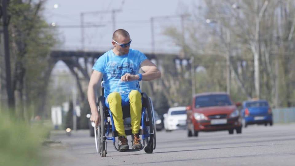 Ein Mann fährt im Rollstuhl auf einer Strasse. Er trägt sportliche Kleidung und schaut auf die Armbanduhr.