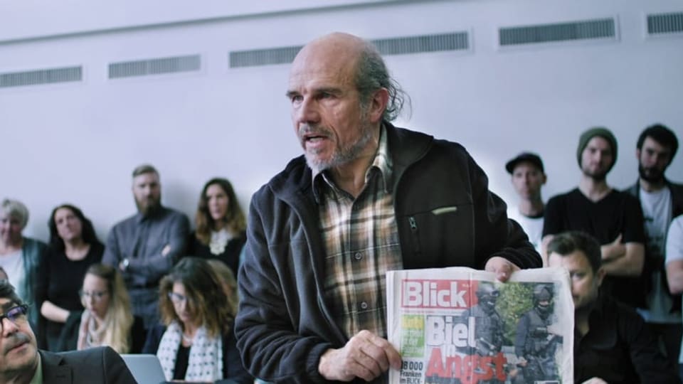 Ein älterer Mann hält eine Schweizer Tageszeitung vor Publikum in die Höhe