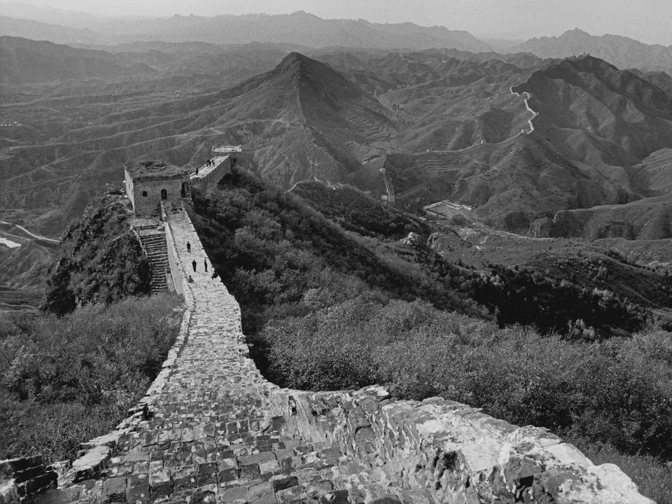 Schwarz-Weiss-Foto einer bergigen Landschaft, durch die sich eine Mauer zieht. 