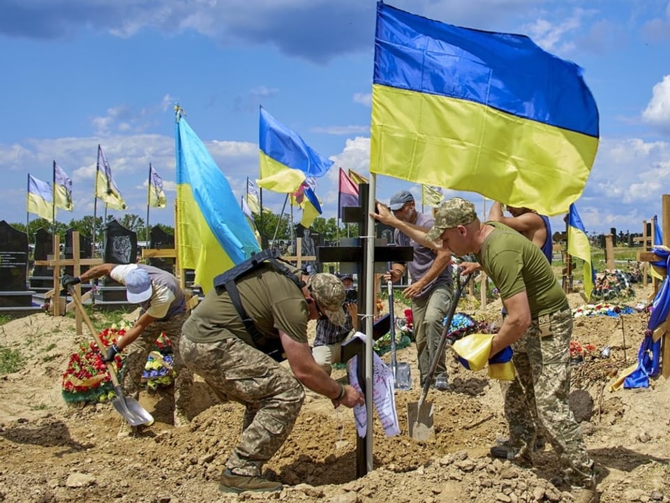 Soldaten heben Gräber aus und stellen ukrainische Flaggen daneben auf. Im Hintergrund wehen schon mehrere Fahnen.