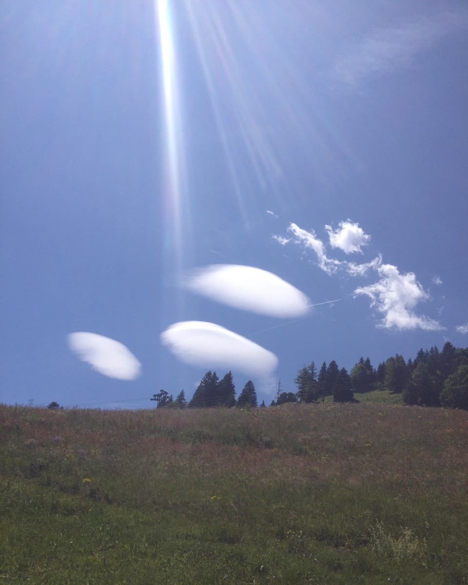 Über einer Wiesen und einem Wald hängen drei mandelförmige Wolken. Die Wolken sehen aus wie drei kleine UFOs.