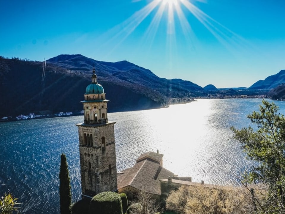 Morcote/TI Blick über Kirche hangabwärts über See mit wolkenlosem Himmel.