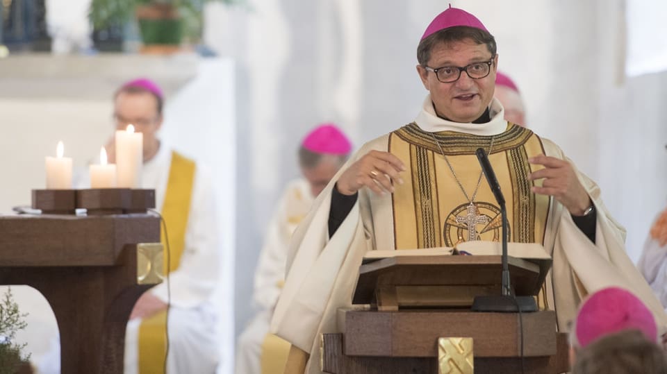 Bischof in weissem gewand und violetter Kappe steht an Holz-Rednerpult, Mund offen, Hände erhoben.