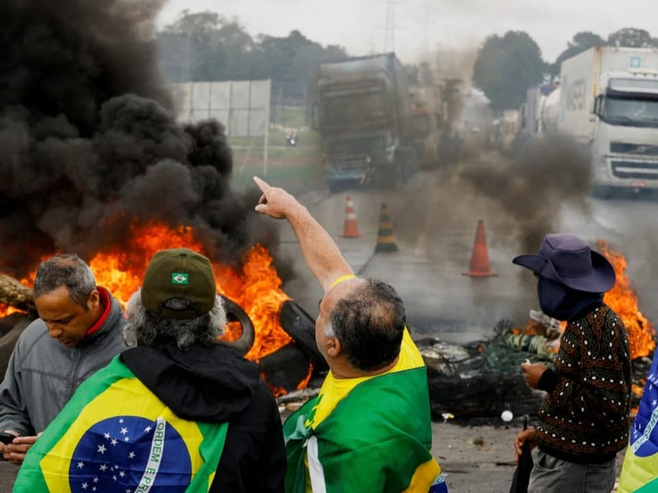Brennende Reifen auf der Strasse. Davor Menschen am protestieren mit grün-gelb-blauen Flaggen, die für Brasilien stehen.