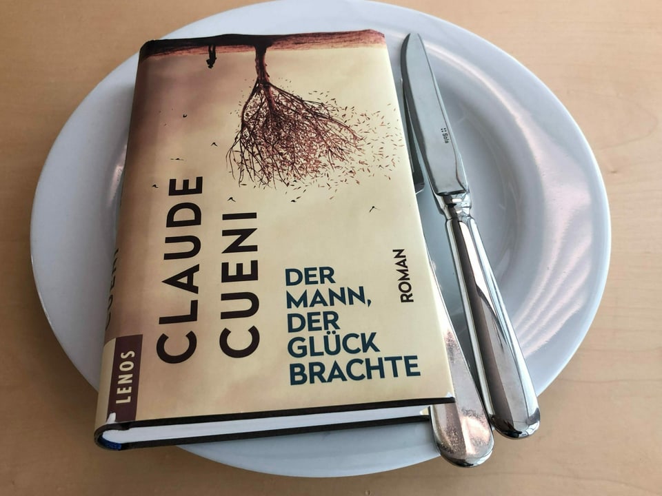 Der Roman «Der Mann, der Glück brachte» liegt auf einem weissen Teller. Messer und Gabel auf der rechten Seite sind neben das Buch gelegt. 