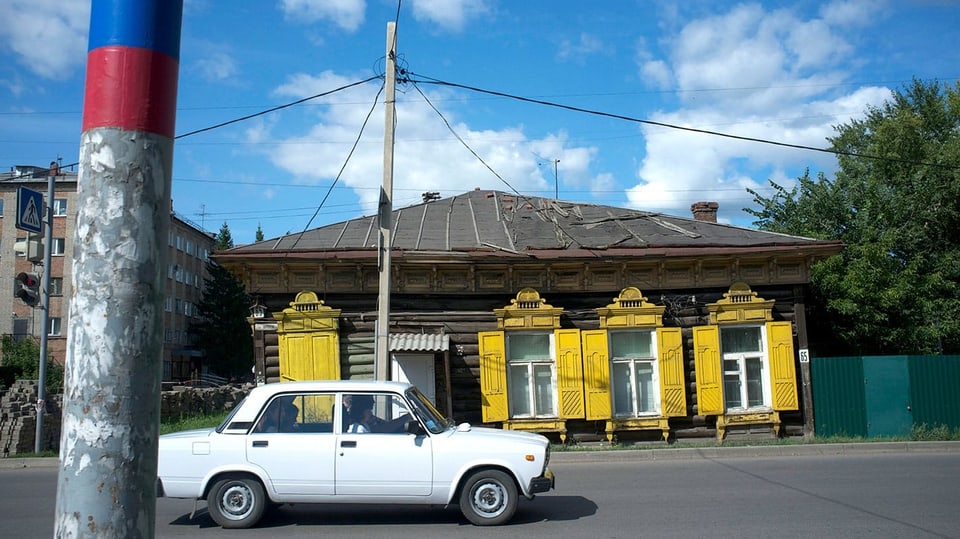 Ein weisser Lada fährt an einem Holzhaus mit gelben Läden vorbei.