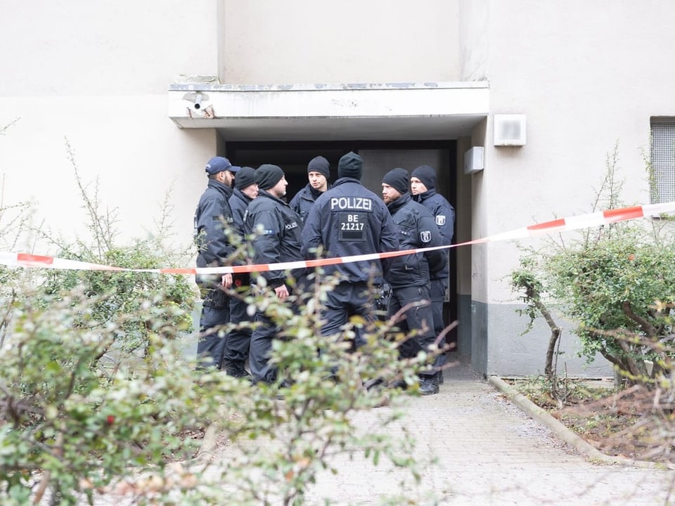 Sieben uniformierte Polizisten stehen hinter rot-weissem Absperrband am Eingang eines Mehrfamilienhauses in Berlin. 