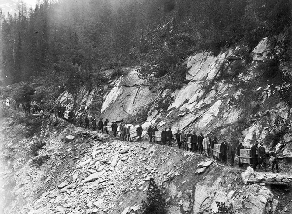 Eine lange Kolonne von Menschen, die jeweils zu zweit eine Holzkiste tragen, windet sich an einem steilen Abhang vorbei - links der Berg, rechts der Abgrund.
