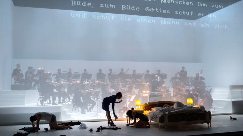 Bühnenbild: Bett steht vor Gazevorhang, dahinter spielt das Orchester.