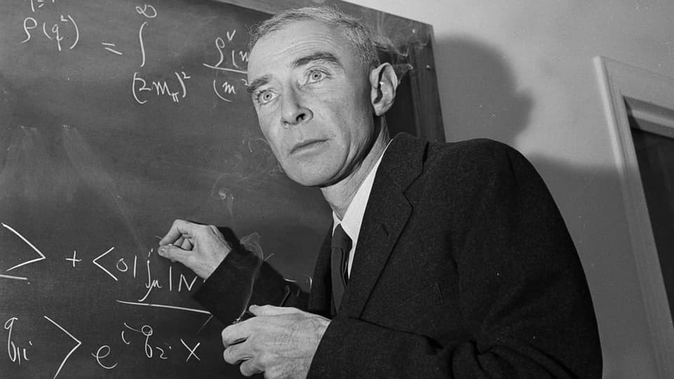 Robert Oppenheimer steht vor einer Wandtafel und schreibt mit Kreide Formeln auf.