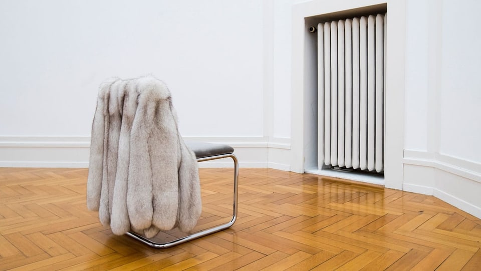 Ein leerer Stuhl mit einem Pelzmantel blickt in Richtung einer Wand mit einem Heizkörper.