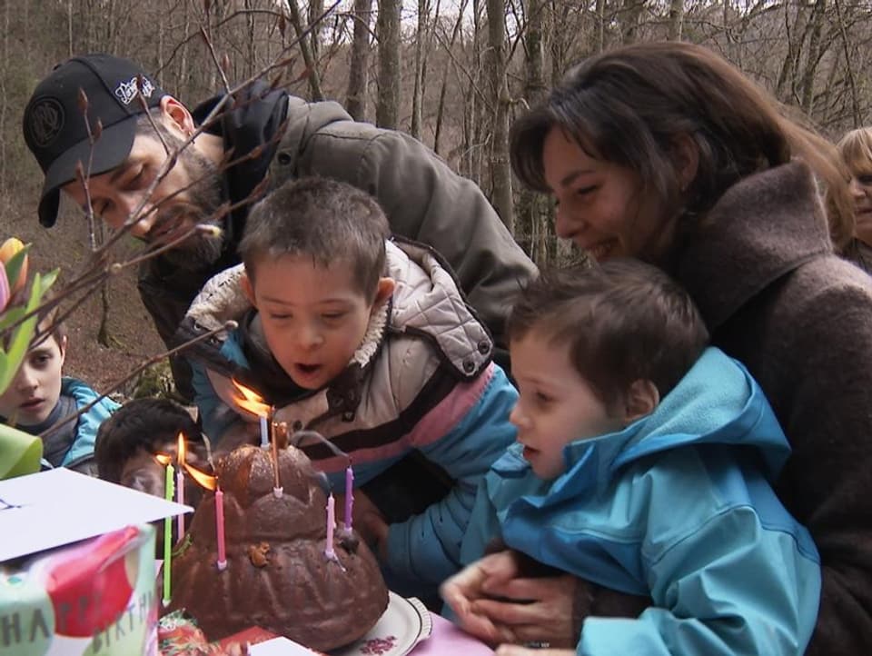 Eine Gruppe von Erwachsenen und Kindern steht vor einem Kuchen. Ein Junge pustet Kerzen auf dem Kuchen aus.