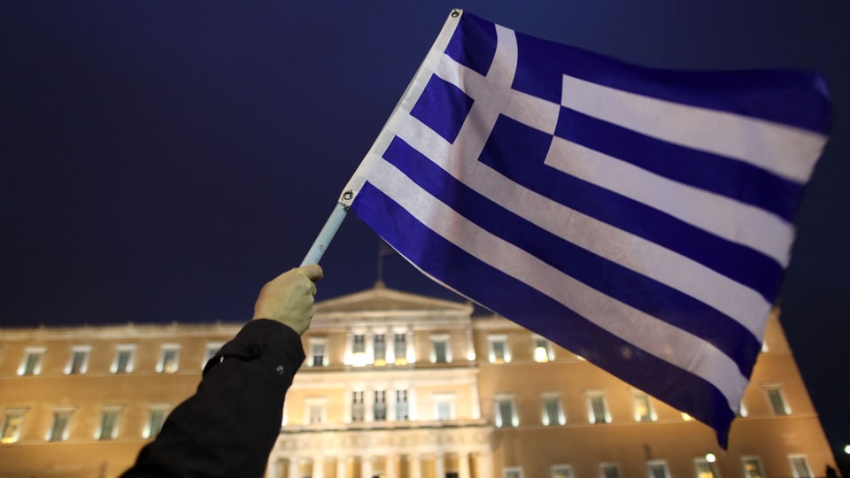 Hand, griechische Fahne haltend, im Hintergrund ist das griechische Parlamentsgebäude sichtbar.