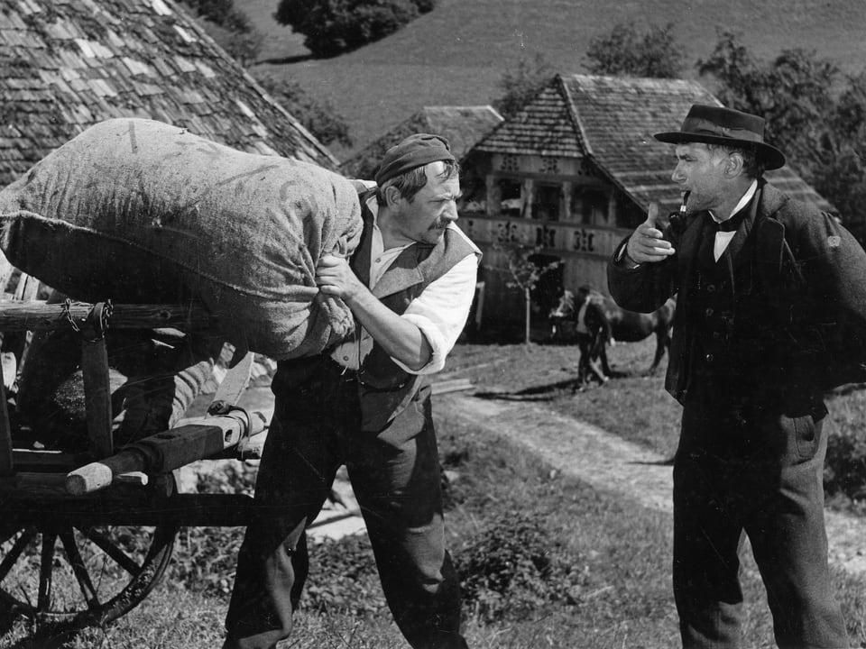 Ein Mann hievt einen Jutesack von einem Wagen. Ein zweiter steht neben ihm und spricht mit ihm während er Pfeiffe raucht. Im Hintergrund ist ein Bauernhaus zu sehen.