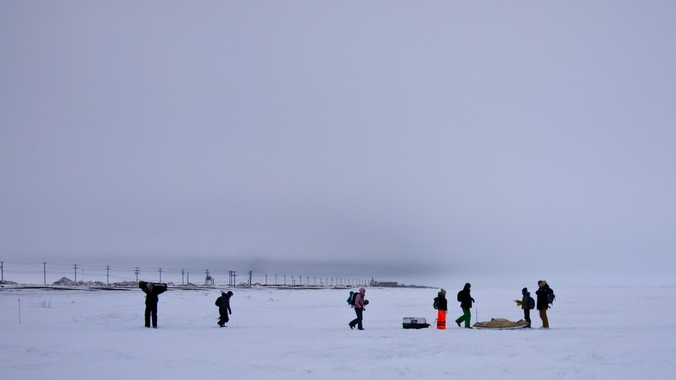 Menschen mit einem Schlitten im Schnee. Im Hintergrund sieht man die Stromleitungen der Küstenstrasse und das Kraftwerk. Eine dunkle Wolke zeigt Barrow an. 