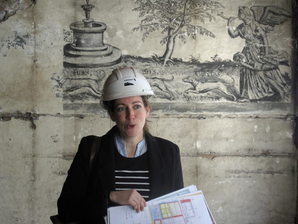 Eine Frau mit Helm steht vor einem Wandgemälde.