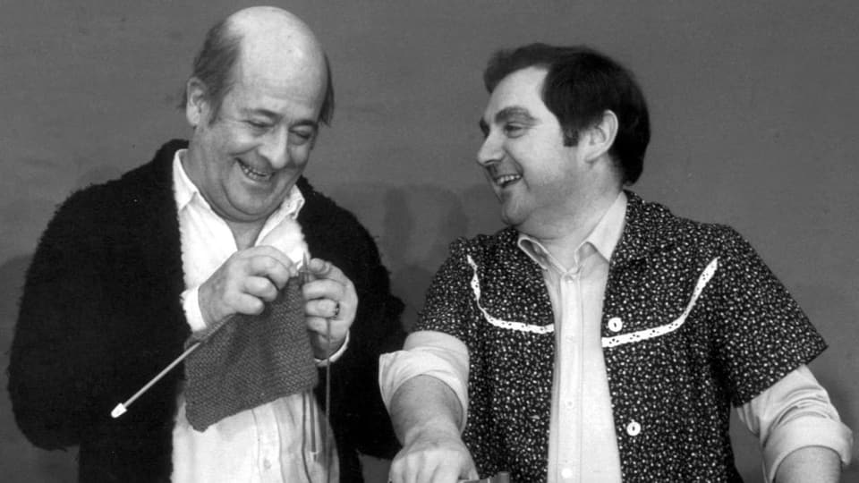 schwarzweiss-Foto zweier Männer, der linke häkelt, der rechte hält ein Bügeleisen. Beide lachen.