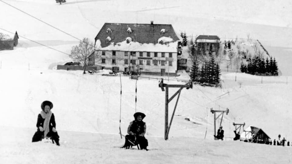 schwarzweiss Foto links oben eine Frau auf dem Schlitten, hinter ihr ein Mann. Beide an einem Bügel des Skilifts.