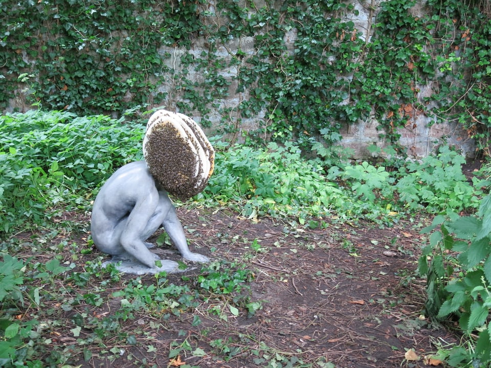 Im Garten sitzt menschliche Skulptur, der Kopf ist ein Bienenstock mit lebenden Bienen drauf. 