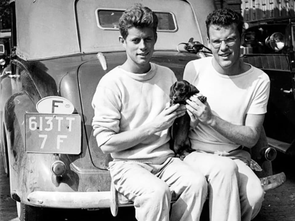 Zwei junge Männer sitzen auf der Stossstange eines Autos und halten einen Hund