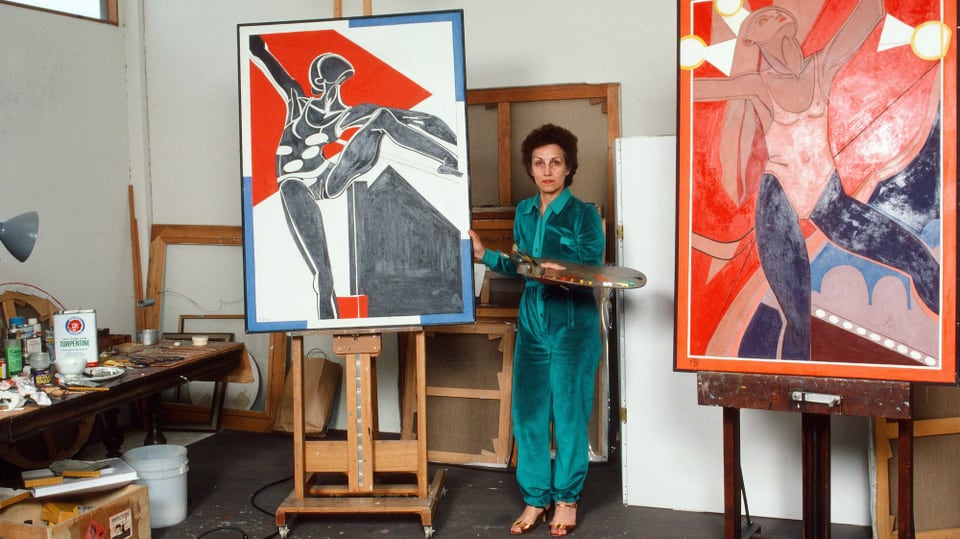 Eine Frau mittleren Alters steht in einem Künstleratelier zwischen zwei Gemälden.