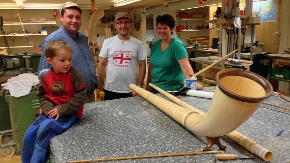 Ein Bub sitzt auf einem Werkstatt-Tisch mit einem Alphorn. Hinter dem Tisch posieren zwei Männer und eine Frau.
