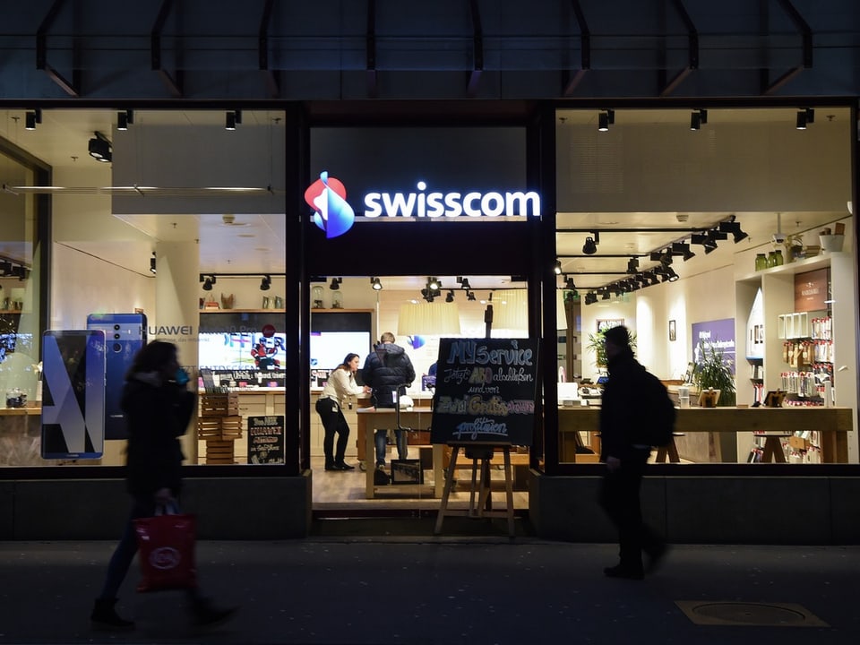 Personen laufen bei Dunkelheit vor einem Swisscom-Shop vorbei. Im Shop bedient eine Verkäuferin einen Kunden.