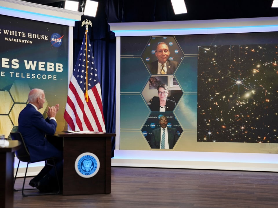 Links im Vordergrund sitzt US-Präsident Joe Biden vor einer Videowand rechts im Bild, auf der drei Personen und das neue Bild des Weltraumteleskops zu sehen sind.