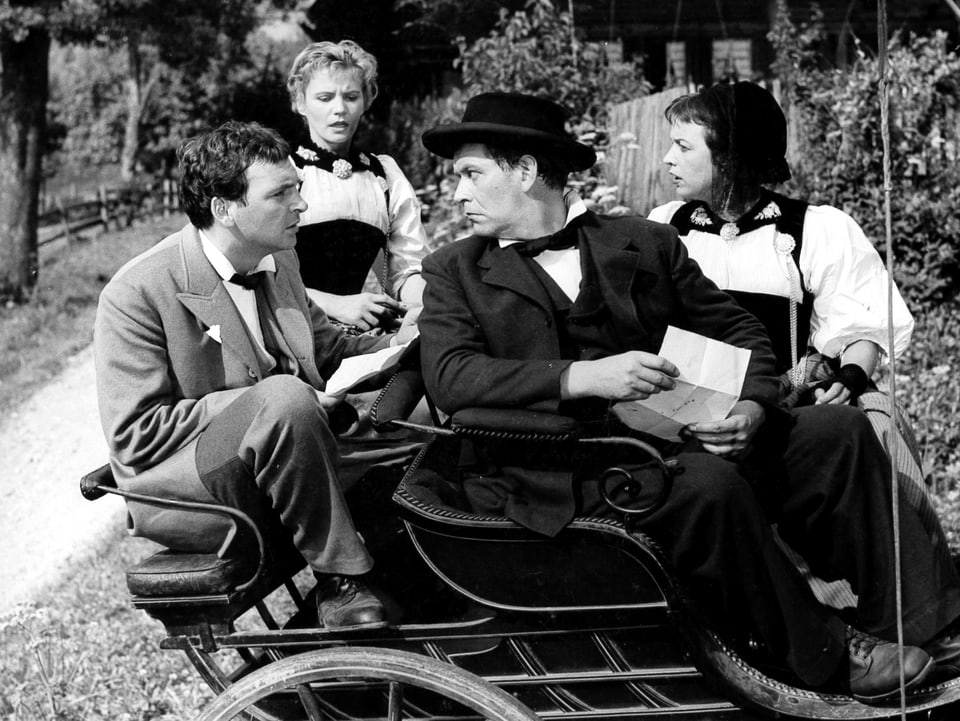 Zwei Männer und eine Frau sitzen auf einer kleinen Kutsche. Eine zweite Frau steht daneben und steigt auch zu. Die beiden Männer blicken sich an. Der Vordere hält ein Blatt Papier in der Hand.