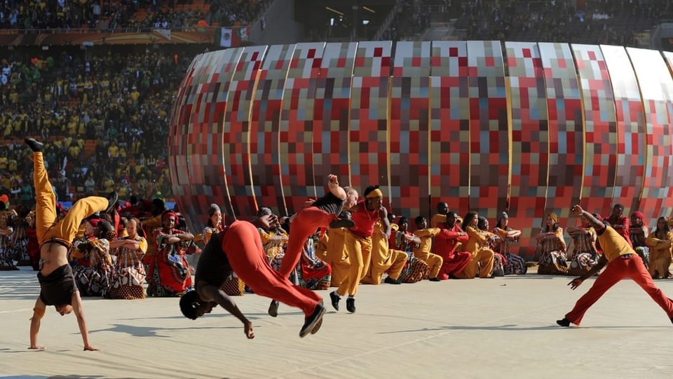 Bei der Eröffnungszeremonie tanzen farbig gekleidete Menschen auf der Bühne, während Tausende im Stadion zuschauen.