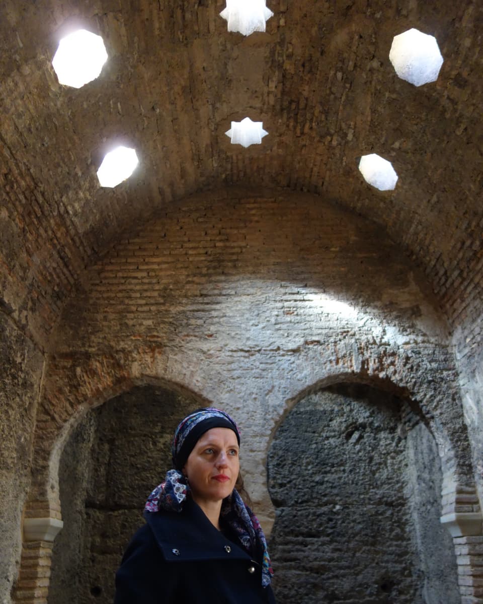 Frau mit Tuch um den Kopf gebunden steht in altem, steinernen Raum. Von oben Licht, achteckige und sternförmige Fenster