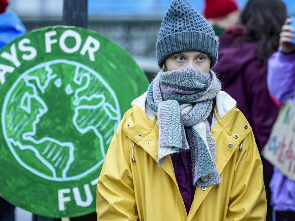 Greta Thunberg steht mit gelber Regenjacke vor einem grünen Schild.