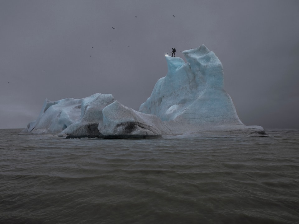 Ein Eisberg im Meer, auf der Spitze steht ein Mann mit einem Flammenwerfer.